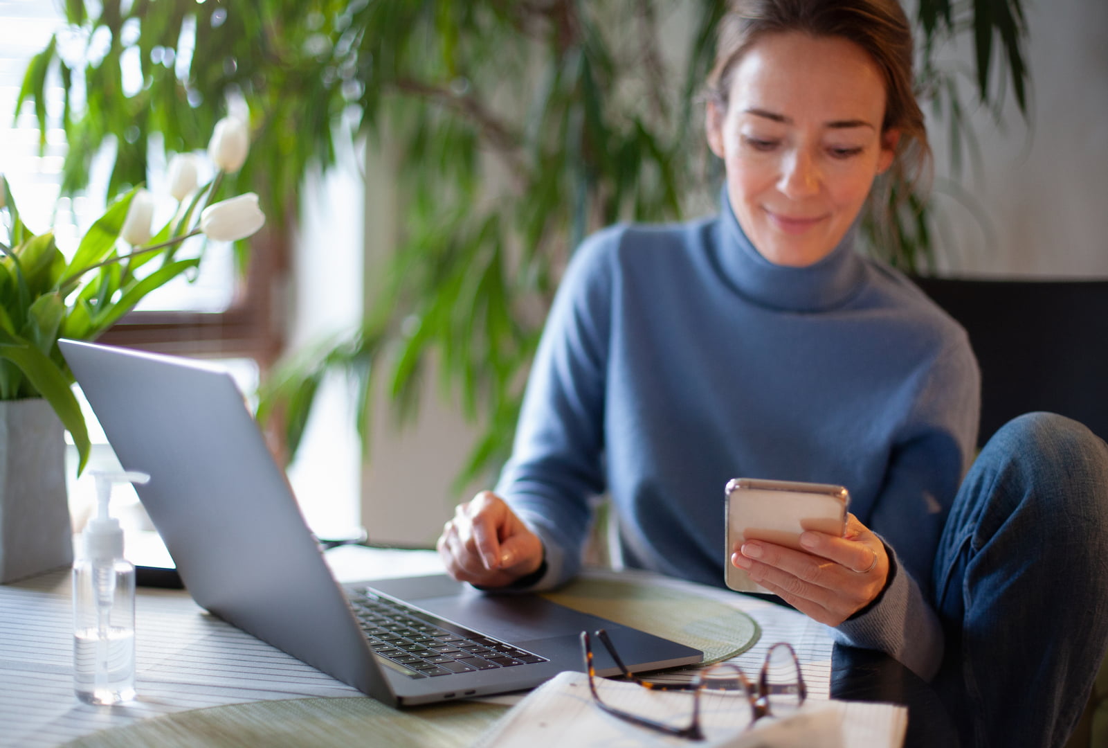 En ung kvinne sitter med laptopen hennes og ser på mobilen