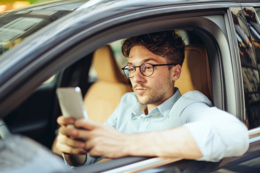 Ung mann med mørkt hår og briller sitter i en bil og ser på en mobiltelefon